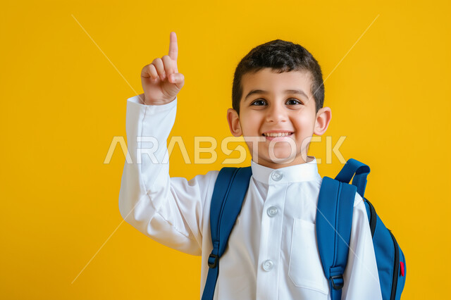 التفاعل والمشاركة داخل الفصل الدراسي، بورتريه مقرب لطالب عربي خليجي سعودي يرتدي الثوب التقليدي وحقيبة الظهر يرفع يده للأعلى للإجابة، التربية والتعليم بالمناهج المقررة للطلاب في المملكة العربية السعودية، خلفية صفراء