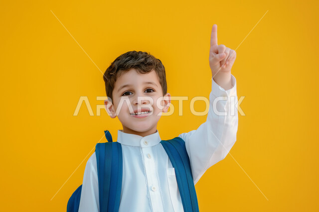 التفاعل والمشاركة داخل الفصل الدراسي، بورتريه مقرب لطالب عربي خليجي سعودي يرتدي الثوب التقليدي وحقيبة الظهر يرفع يده للأعلى للإجابة، التربية والتعليم بالمناهج المقررة للطلاب في المملكة العربية السعودية، خلفية صفراء