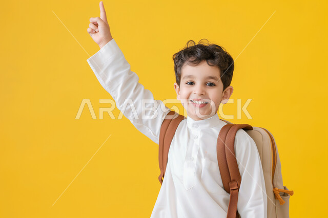 التفاعل والمشاركة داخل الفصل الدراسي، بورتريه مقرب لطالب عربي خليجي سعودي يرتدي الثوب التقليدي وحقيبة الظهر يرفع يده للأعلى للإجابة، التربية والتعليم بالمناهج المقررة للطلاب في المملكة العربية السعودية، خلفية صفراء