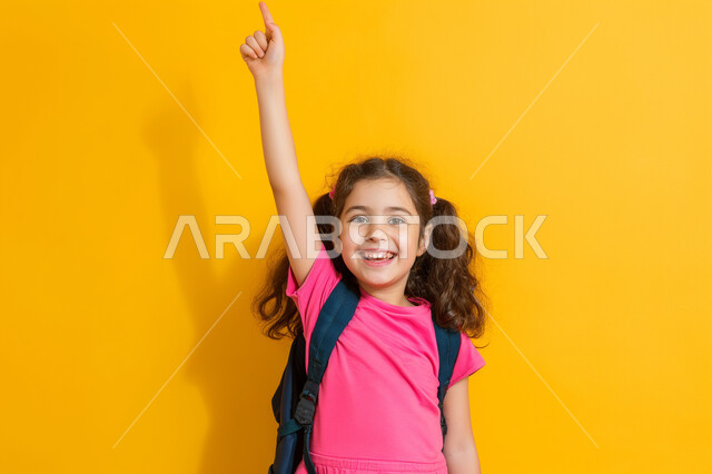 المشاركة والتفاعل داخل الفصل الدراسي، بورتريه مقرب لطالبة عربية خليجية سعودية ترتدي حقيبة الظهر ترفع يدها للأعلى للإجابة، التربية والتعليم بالمناهج المقررة للطلاب في المملكة العربية السعودية، خلفية صفراء