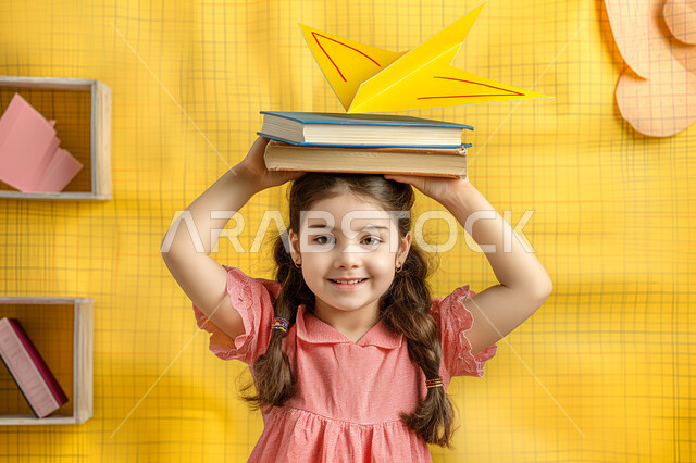 النظر الى الكاميرا بتعابير وايماءات السرور والسعادة، حب القراءة والثقافة والإستطلاع، بورتريه مقرب لطالبة عربية خليجية سعودية مبتسمة تحمل بيدها مجموعة من الكتب، مراجعة الدروس اليومية، التعليم والمدارس في المملكة العربية السعودية، خلفية صفراء