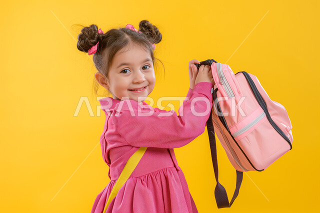 النشاط الصباحي والجد والاجتهاد، بورتريه لفتاةعربية خليجية سعودية ترتدي الحقيبة المدرسية تقوم بالذهاب الى المدرسة، بدء الدوام الصباحي في مدارس المملكة العربية السعودية، صورة الجسم بالكامل، خلفية صفراء