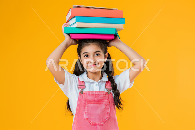 النظر الى الكاميرا بتعابير وايماءات السرور والسعادة، حب القراءة والثقافة والإستطلاع، بورتريه مقرب لطالبة عربية خليجية سعودية مبتسمة تحمل بيدها مجموعة من الكتب، مراجعة الدروس اليومية، التعليم والمدارس في المملكة العربية السعودية، خلفية صفراء