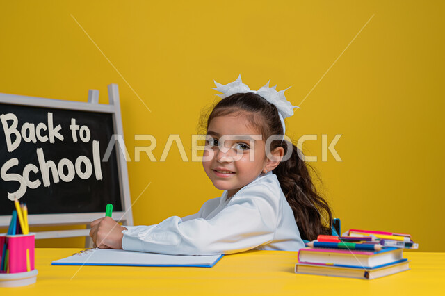 Back to school, education in Saudi Arabia schools, close-up portrait of smiling Saudi Arabian Gulf girl sitting at study table reviewing lessons and doing homework, school supplies and books, diligent student looking at camera with happy expression, yellow background