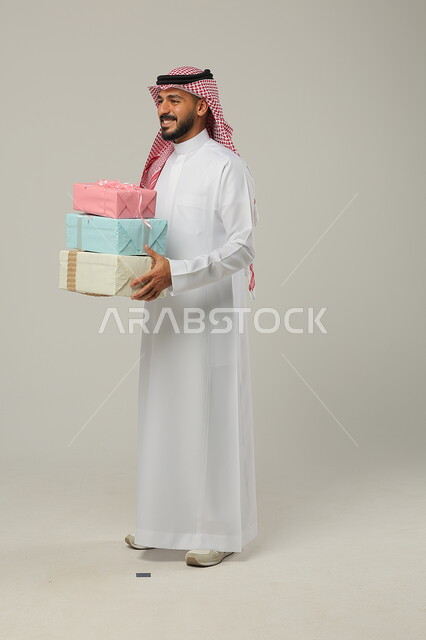 المباركات والتهنئات بالنجاح والأفراح, تقديم الهدية في مناسبات الأهل والأصدقاء المقربة, الاحتفال بالأعياد السعيدة, بورتريه لرجل عربي خليجي سعودي مبتسم يرتدي الثوب التقليدي يحمل بيديه علب هدايا مغلفة بألوان أنيقة, الجسم بالطول الكامل, خلفية رمادية