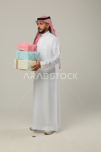 المباركات والتهنئات بالنجاح والأفراح, تقديم الهدية في مناسبات الأهل والأصدقاء المقربة, الاحتفال بالأعياد السعيدة, بورتريه لرجل عربي خليجي سعودي مبتسم يرتدي الثوب التقليدي يحمل بيديه علب هدايا مغلفة بألوان أنيقة, الجسم بالطول الكامل, خلفية رمادية