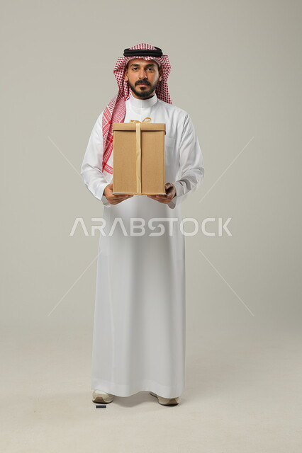 أعياد الميلاد والمباركات والأفراح, تقديم الهدايا بالمناسبات العائلية السعيدة, زيادة روابط المحبة بالإهداء والتقدير, بورتريه لشاب عربي خليجي سعودي وسيم يرتدي الثوب التقليدي يمسك بيده علبة هدية ملفوفة بشريط أنيق, الجسم بالطول الكامل, خلفية رمادية