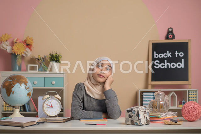 Thinking and absent-mindedness, looking somewhere with frowning and boredom, feeling and sensing boredom and exhaustion from solving daily homework, a Saudi Arabian Gulf girl sitting at the study table with expressions of waiting for something