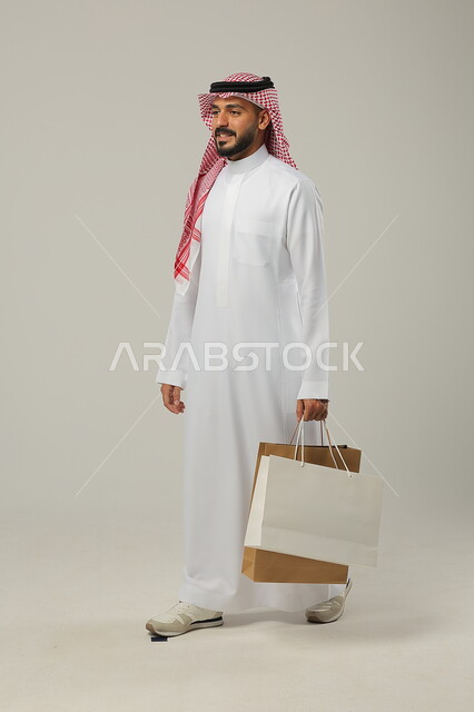 التبضع وأخذ الحاجات اللازمة والضرورية, المشتريات التجارية والهدايا, مواسم التخفيضات في السعودية, الاستفادة من الحسومات والتصفيات, بورتريه لشاب عربي خليجي سعودي يرتدي الثوب التقليدي والشماغ يمسك بيده أكياس ورقية, متعة التسوق في المملكة, جسم بالطول الكامل, خلفية رمادية