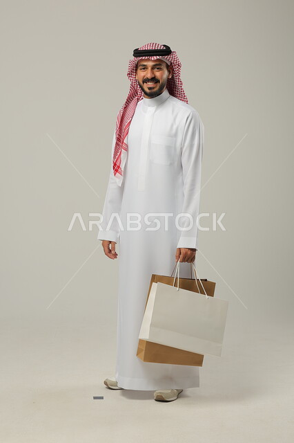 التبضع وأخذ الحاجات اللازمة والضرورية, المشتريات التجارية والهدايا, مواسم التخفيضات في السعودية, الاستفادة من الحسومات والتصفيات, بورتريه لشاب عربي خليجي سعودي يرتدي الثوب التقليدي والشماغ يمسك بيده أكياس ورقية, متعة التسوق في المملكة, جسم بالطول الكامل, خلفية رمادية