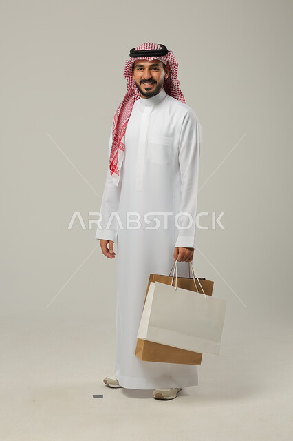 التبضع وأخذ الحاجات اللازمة والضرورية, المشتريات التجارية والهدايا, مواسم التخفيضات في السعودية, الاستفادة من الحسومات والتصفيات, بورتريه لشاب عربي خليجي سعودي يرتدي الثوب التقليدي والشماغ يمسك بيده أكياس ورقية, متعة التسوق في المملكة, جسم بالطول الكامل, خلفية رمادية