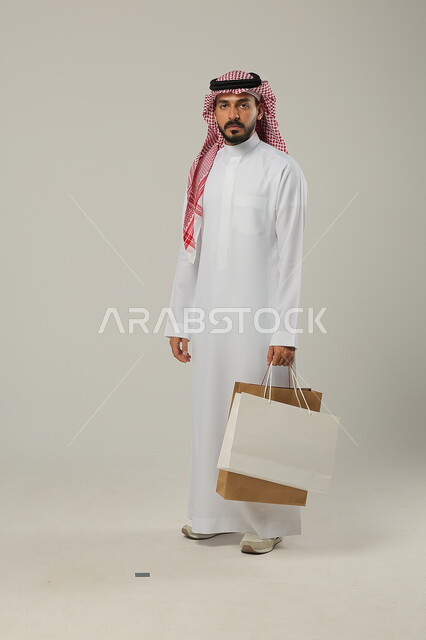 التبضع وأخذ الحاجات اللازمة والضرورية, المشتريات التجارية والهدايا, مواسم التخفيضات في السعودية, الاستفادة من الحسومات والتصفيات, بورتريه لشاب عربي خليجي سعودي يرتدي الثوب التقليدي والشماغ يمسك بيده أكياس ورقية, متعة التسوق في المملكة, جسم بالطول الكامل, خلفية رمادية