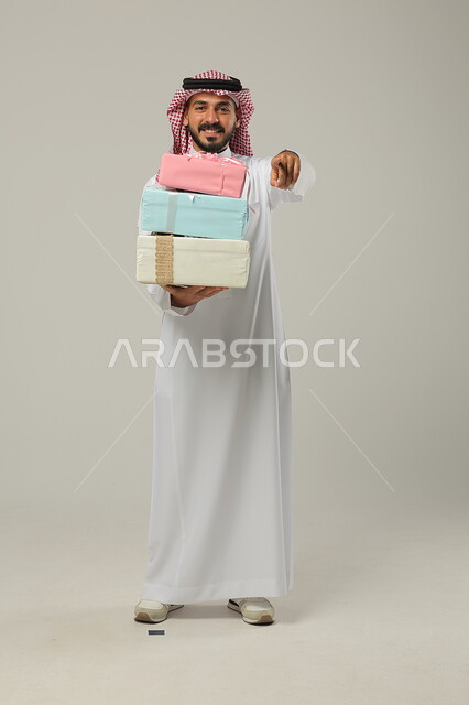 التهنئات والمباركات بالنجاح والأفراح, الإشارة إلى الهدايا بإيماءات المفاجأة، تقديم الهدية في مناسبات الأهل والأصدقاء المقربة, بورتريه لرجل عربي خليجي سعودي مبتسم يرتدي الثوب التقليدي يحمل بيديه علب مغلفة بألوان أنيقة, الجسم بالطول الكامل, خلفية رمادية