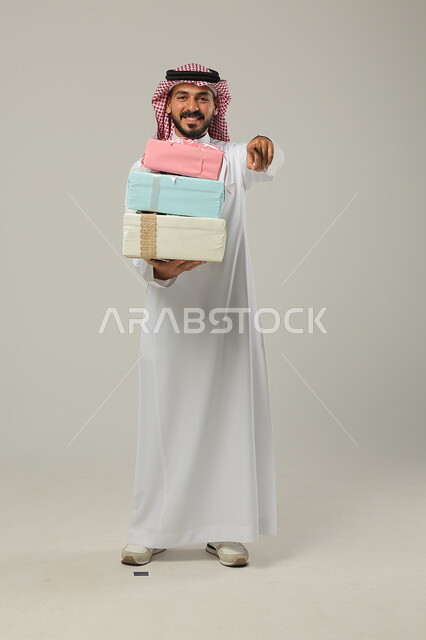 التهنئات والمباركات بالنجاح والأفراح, الإشارة إلى الهدايا بإيماءات المفاجأة، تقديم الهدية في مناسبات الأهل والأصدقاء المقربة, بورتريه لرجل عربي خليجي سعودي مبتسم يرتدي الثوب التقليدي يحمل بيديه علب مغلفة بألوان أنيقة, الجسم بالطول الكامل, خلفية رمادية
