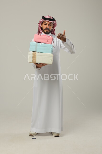 التهنئات والمباركات بالنجاح والأفراح, الإشارة إلى الهدايا بإيماءات المفاجأة، تقديم الهدية في مناسبات الأهل والأصدقاء المقربة, بورتريه لرجل عربي خليجي سعودي مبتسم يرتدي الثوب التقليدي يحمل بيديه علب مغلفة بألوان أنيقة, الجسم بالطول الكامل, خلفية رمادية