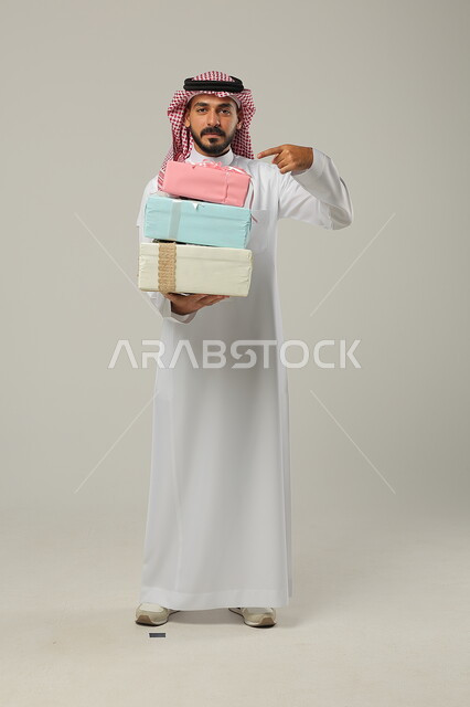 التهنئات والمباركات بالنجاح والأفراح, الإشارة إلى الهدايا بإيماءات المفاجأة، تقديم الهدية في مناسبات الأهل والأصدقاء المقربة, بورتريه لرجل عربي خليجي سعودي مبتسم يرتدي الثوب التقليدي يحمل بيديه علب مغلفة بألوان أنيقة, الجسم بالطول الكامل, خلفية رمادية