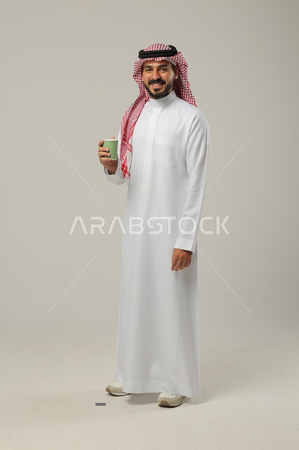 Looking at the camera and enjoying a favorite drink, sipping a delicious hot drink, portrait of a Saudi Gulf Arab young man wearing a traditional dress and holding a cup of coffee in his hand, authentic fine Arabic coffee, enjoying drinks in cafes and ready-to-eat restaurants, full-length body, gray background