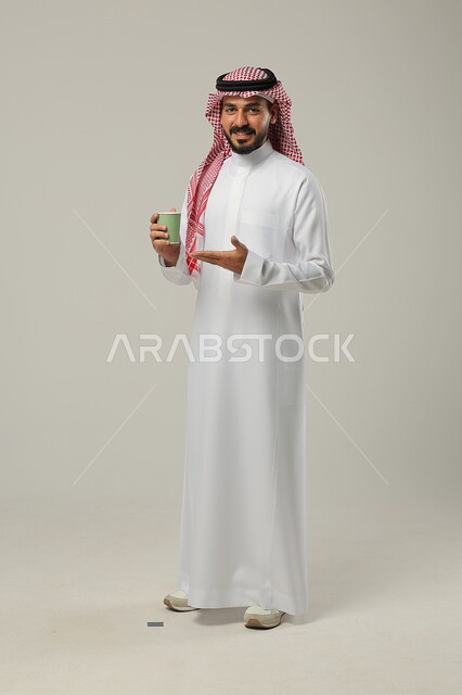 Enjoying favorite drinks, sipping a delicious hot drink, portrait of a Saudi Gulf Arab young man wearing a traditional dress raising his hand up to point to a cup of coffee, authentic luxurious Arabic coffee, enjoying drinks from cafes and ready-to-eat restaurants, full-length body, gray background