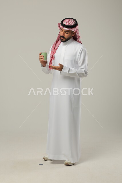 احتساء مشروب ساخن لذيذ, الاستمتاع بتناول المشروبات المفضلة, بورتريه لشاب عربي خليجي سعودي يرتدي الثوب التقليدي يحمل بيده كوب من القهوة, البن العربي الفاخر, التلذذ بمشاريب الكافيهات والمطاعم الجاهزة، الجسم بالطول الكامل، خلفية رمادية