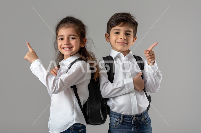 الإستعداد للذهاب إلى المدرسة، الإشارة الى شيء ما، بورتريه لطالبين عربيين خليجيين سعوديين يرتديان الزي المدرسي يحملان الحقيبة المدرسية ينظران الى الكاميرا بتعابير الحماس، مفهوم التعليم الدراسي في السعودية، خلفية رمادية