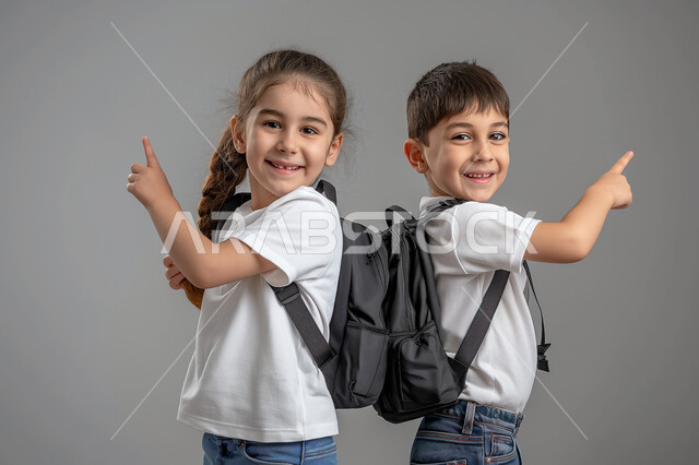 الإستعداد للذهاب إلى المدرسة، الإشارة الى شيء ما، بورتريه لطالبين عربيين خليجيين سعوديين يرتديان الزي المدرسي يحملان الحقيبة المدرسية ينظران الى الكاميرا بتعابير الحماس، مفهوم التعليم الدراسي في السعودية، خلفية رمادية