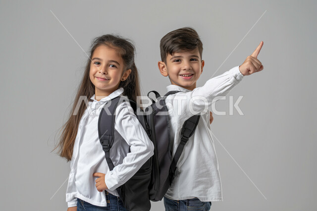 الإستعداد للذهاب إلى المدرسة، الإشارة الى شيء ما، بورتريه لطالبين عربيين خليجيين سعوديين يرتديان الزي المدرسي يحملان الحقيبة المدرسية ينظران الى الكاميرا بتعابير الحماس، مفهوم التعليم الدراسي في السعودية، خلفية رمادية