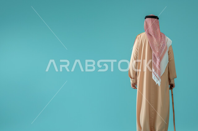 الاعتماد على الوسائل المساعدة في المشي والتنقل، التقدم بالعمر والهيبة والوقار، مفهوم الرجولة العربية, بورتريه من الخلف لرجل كبير بالسن عربي خليجي سعودي يرتدي الثوب التقليدي ويضع الشماغ على رأسه يستند على عكازه, خلفية ملونة