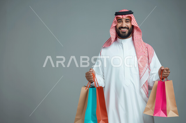 التنزيلات والحسومات الموسمية، الاستفادة من العروض والتخفيضات, بورتريه لرجل عربي خليجي سعودي يرتدي الثوب التقليدي والشماغ يحمل بيده أكياس ورقية ملونة وينظر إلى الكاميرا بإيماءات السرور والسعادة، التسوق واقتناء المستلزمات والهدايا, خلفية رمادية