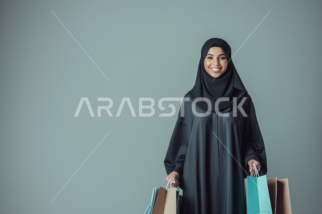 التسوق واقتناء المستلزمات والهدايا, النظر إلى الكاميرا بإيماءات السرور والسعادة، العروض والتخفيضات على كافة الأصناف, الحسومات والتنزيلات الموسمية، بورتريه لامرأة عربية خليجية سعودية  محجبة ترتدي العباءة السوداء تحمل بيدها أكياس ورقية، خلفية رمادية