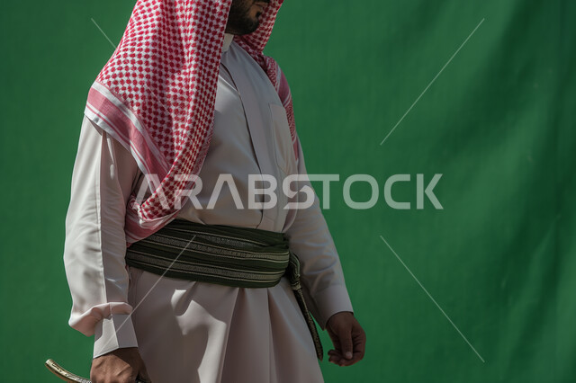 ذكرى يوم التأسيس السعودي, ارتداء أزياء شعبية تقليدية, إحياء تراث المملكة, الاحتفالات بالمناسبات الوطنية, الفخر والاعتزاز بالانتماء الوطني, بورتريه مقرب لزي تراثي لرجل عربي خليجي سعودي يمسك بيده السيف, تأسيس الدولة السعودية الأولى 1727م، خلفية خضراء