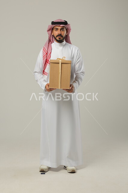 Preparing to give gifts, holidays, occasions and happy surprises, a portrait of a Saudi Gulf Arab man wearing the traditional Saudi thobe and shemagh, full body portrait, gray background