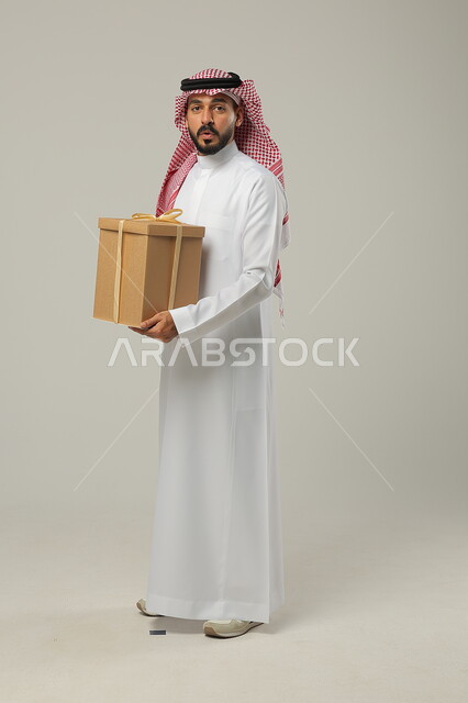  تقديم الهدايا بالمناسبات، أعياد ومفاجأت سعيدة، بورتريه من الجانب لرجل عربي سعودي خليجي يرتدي الثوب و الشماغ السعودي التقليدي ، صورة لكامل الجسم، خلفية رمادية 