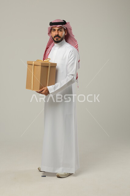 النظر إلى الكاميرا، تقديم الهدايا بالمناسبات، أعياد ومفاجأت سعيدة، بورتريه من الجانب لرجل عربي سعودي خليجي يرتدي الثوب و الشماغ السعودي التقليدي ، صورة لكامل الجسم، خلفية رمادية 