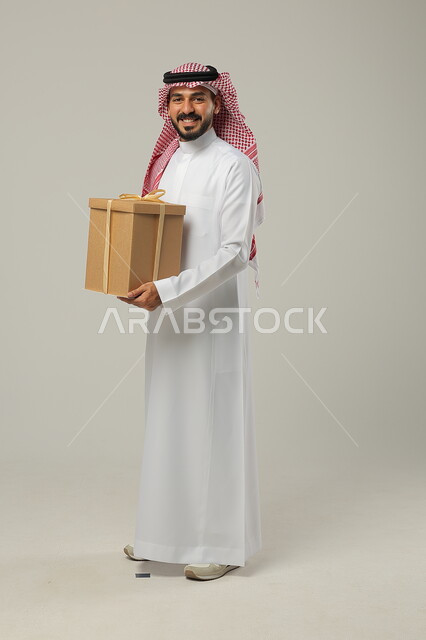 مناسبات واعياد سعيدة، النظر إلى الكاميرا، تقديم الهدايا بالمناسبات، بورتريه من الجانب لرجل عربي سعودي خليجي يرتدي الثوب و الشماغ السعودي التقليدي ، صورة لكامل الجسم، خلفية رمادية 