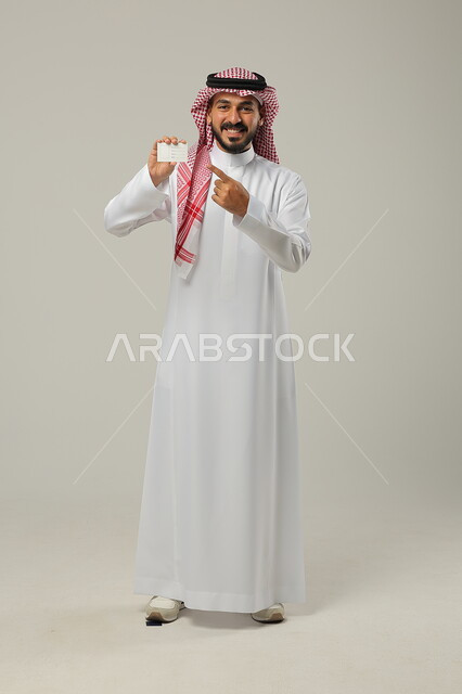  الوقوف باستقامة وثقة، النظر الى الكاميرا بإيماءات السرور و الرضا، بورتريه لرجل عربي خليجي سعودي مبتسم يرتدي الثوب السعودي  التقليدي والشماغ و يحمل بيده بطاقة تعريفية، استخدام بطاقات توضيحية، الجسم بالطول الكامل، خلفية رمادية 