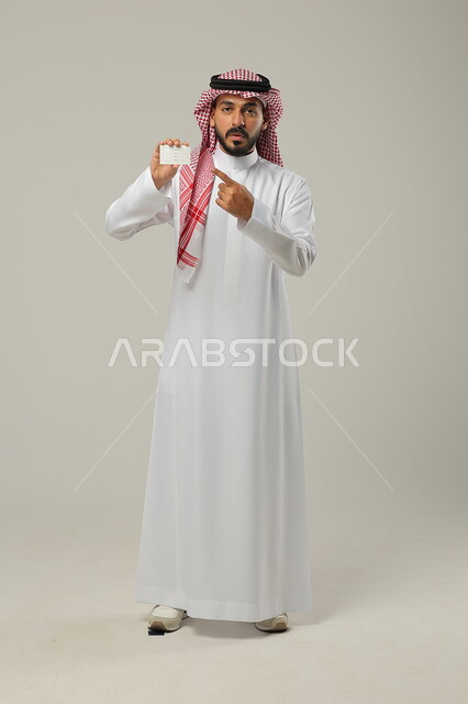  الإشارة الى امر ما، ايماءات الدهشة والتعجب، استخدام بطاقات توضيحية تعريفية، بورتريه لرجل عربي خليجي سعودي يرتدي الشماغ والثوب التقليدي ينظر إلى الكاميرا، صورة الجسم بالكامل، خلفية رماديه 