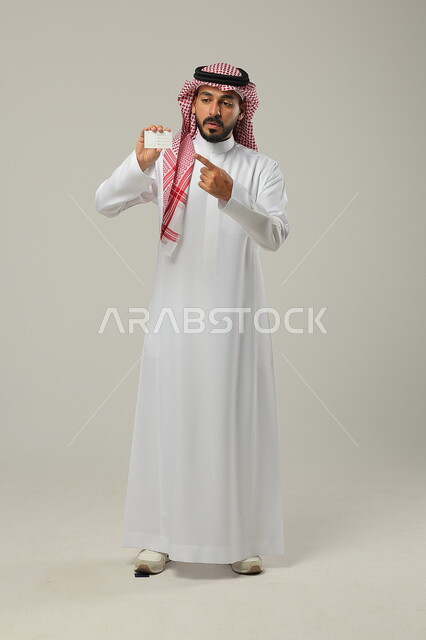 التعابيروالايماءات الدهشة وتوضيح أمرما،  استخدام بطاقة توضيحية، بورتريه لرجل عربي خليجي سعودي يرتدي الثوب السعودي التقليدي والشماغ و يحمل بيده بطاقة تعريفية، الجسم بالطول الكامل، خلفية رمادية 