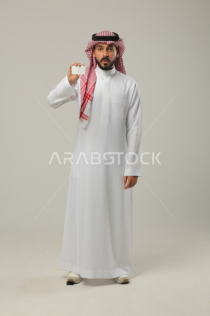 Gestures of shock and astonishment, blank white rectangular card, use of identification cards, portrait of a Saudi Gulf Arab man wearing a shemagh and traditional dress looking at the camera, full body portrait, gray background