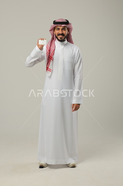 Using an explanatory card, standing upright and confident, looking at the camera with gestures of pleasure and satisfaction, a portrait of a smiling Saudi Gulf Arab man wearing the traditional Saudi thobe and shemagh and holding an identification card in his hand, full length body, gray background.