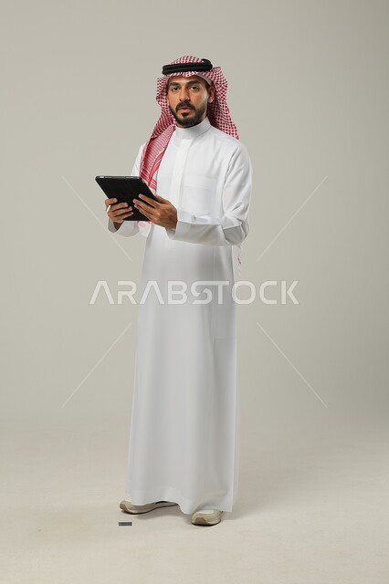E-shopping through programs and applications, a portrait of a Saudi Gulf Arab man wearing traditional clothing and a shemagh and holding a tablet in his hand, gestures of seriousness and firmness, full-length body portrait, gray background