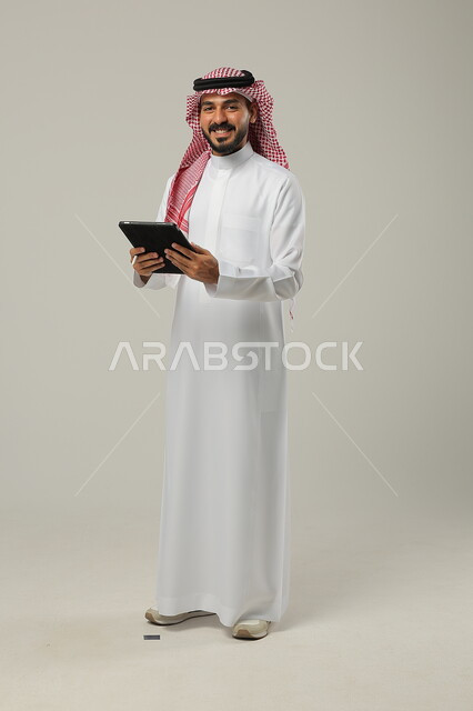 إيماءات السروروالفرح، التسوق الالكتروني عبر البرامج والتطبيقات ، بورتريه لرجل عربي خليجي سعودي يرتدي الزي التقليدي والشماغ ويحمل بيده جهازالتابلت، صورة الجسم بالطول الكامل، خلفية رمادية 