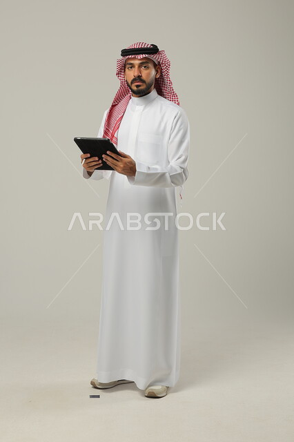 إستخدام اجهزة تقنية حديثة، إيماءات وتعابير الدهشة والصدمة، مفهوم إدارة المشاريع عن بعد، بورتريه لرجل عربي خليجي سعودي يرتدي الشماغ والثوب التقليدي يقف باستقامة ويقوم بإنجاز مهام العمل عبر جهاز التابلت، صورة الجسم بالطول الكامل،خلفية رمادية 