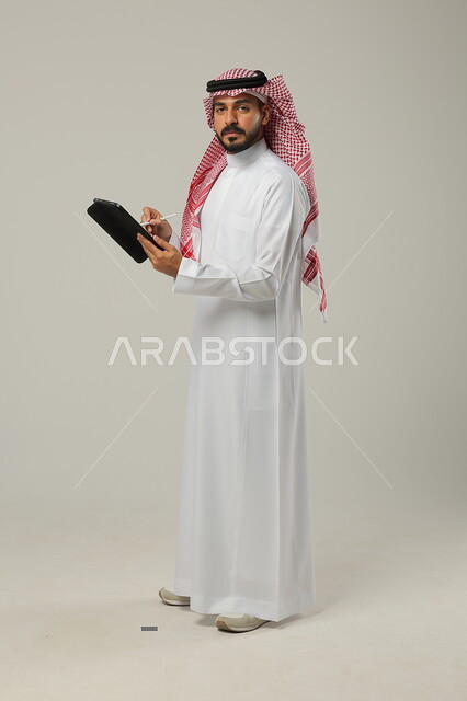 مفهوم إدارة المشاريع عن بعد، إستخدام اجهزة تقنية حديثة، إيماءات وتعابير الدهشة والصدمة، بورتريه لرجل عربي خليجي سعودي يرتدي الشماغ والثوب التقليدي يقف باستقامة ويقوم بإنجاز مهام العمل عبر جهاز التابلت، صورة الجسم بالطول الكامل،خلفية رمادية 