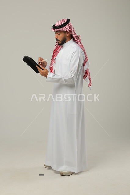  استخدام الأجهزة اللوحية الحديثة المتطورة، ايماءات وتعابير الاندهاش والتركيز، التواصل والدردشة مع الاقارب و الاصدقاء، بورتريه لرجل عربي خليجي سعودي يرتدي الشماغ والثوب التقليدي يمسك بيده جهاز التابلت، خلفية رمادية 