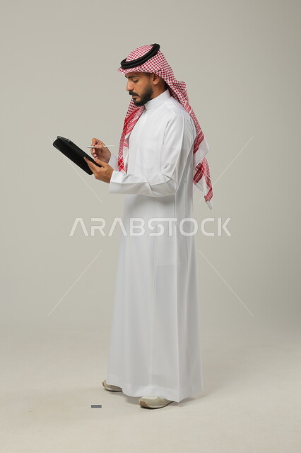إنجازمهام العمل عن بعد، متابعة آخر الأخبارعلى مواقع الانترنت، بورتريه لرجل عربي خليجي سعودي مبتسم يرتدي الزي التقليدي والشماغ يحمل بيده جهاز التابلت، دمج التكنولوجيا والتقنية الحديثة بالحياة العملية، صورة الجسم بالطول الكامل، خلفية رمادية