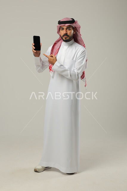  استخدام التكنولوجيا المتطورة، عرض تطبيقات وخدمات الهاتف المحمول، مفهوم التسويق الرقمي الالكتروني، بورتريه لشاب عربي خليجي سعودي مبتسم يرتدي االشماغ ولثوب التقليدي يحمل بيده جوال بشاشة سوداء فارغة ويشير اليه باصبع السبابة، صورة الجسم بالطول الكامل، خلفية رمادية 