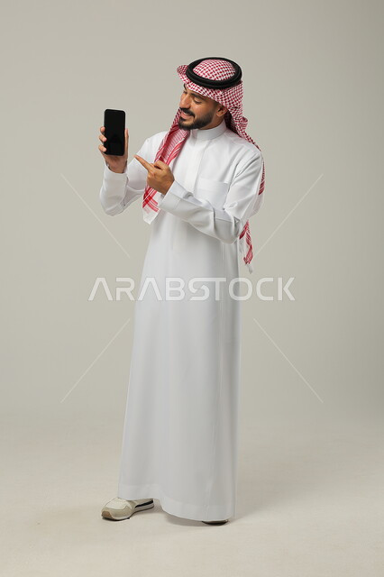  عرض تطبيقات وخدمات الهاتف المحمول، مفهوم التسويق الرقمي الالكتروني، بورتريه لشاب عربي خليجي سعودي مبتسم يرتدي االشماغ ولثوب التقليدي يحمل بيده جوال بشاشة سوداء فارغة ويشير اليه، استخدام التكنولوجيا المتطورة، صورة الجسم بالطول الكامل، خلفية رمادية 