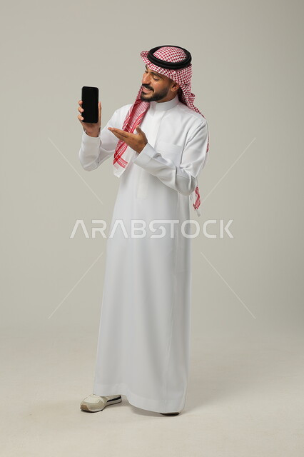 التواصل مع الأهل والأصدقاء، إجراء مكالمات فيديو بإيماءات الفرح والسرور، استخدام التقنيات الحديثة لالتقاط الصور التذكارية، بورتريه لشاب عربي سعودي خليجي مبتسم يرتدي الثوب والشماغ التقليدي يشير إلى شاشة الهاتف المحمول ،  صورة الجسم بالطول الكامل, خلفية رمادية