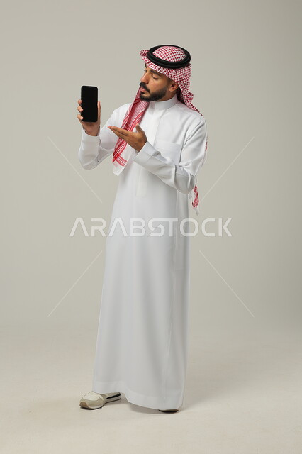  مفهوم التسويق الرقمي الالكتروني، عرض تطبيقات وخدمات الهاتف المحمول، بورتريه لشاب عربي خليجي سعودي يرتدي االشماغ ولثوب التقليدي يحمل بيده جوال بشاشة سوداء فارغة ويشير اليه، استخدام التكنولوجيا المتطورة، صورة الجسم بالطول الكامل، خلفية رمادية 