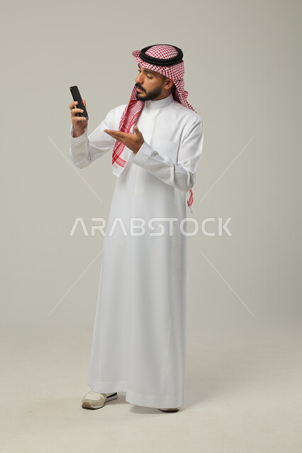  استخدام التقنيات الحديثة لالتقاط الصورالصورالتذكارية، التواصل مع الأهل والأصدقاء، إجراء مكالمات فيديو بإيماءات الانهاش والتساؤل، بورتريه لشاب عربي سعودي خليجي يرتدي الثوب والشماغ التقليدي يشير إلى شاشة الهاتف المحمول ،  صورة الجسم بالطول الكامل، خلفية رمادية